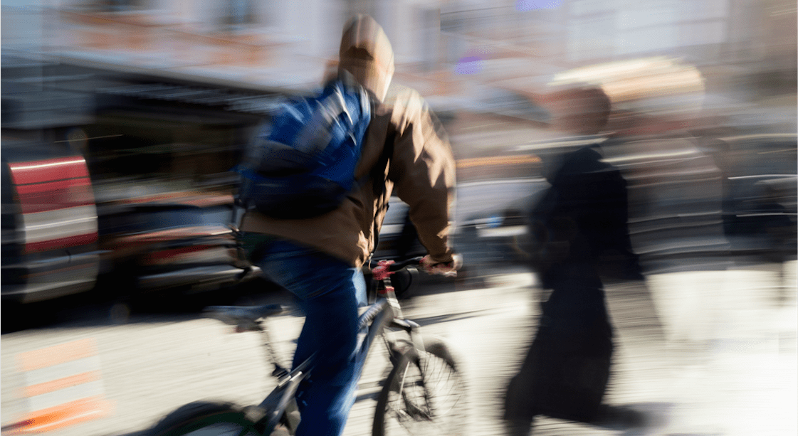 cyklista, před ním přebíhající chodec, jde o rozmazané foto jako by ve velké rychlosti